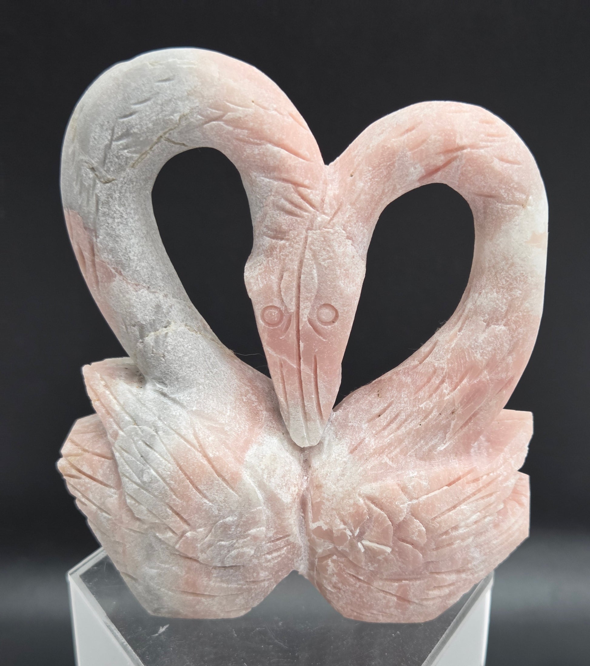 A close-up of a sculpted decorative object featuring two intertwined looped forms resembling a stylized bird or abstract sculpture, mounted on a clear acrylic stand against a dark background.