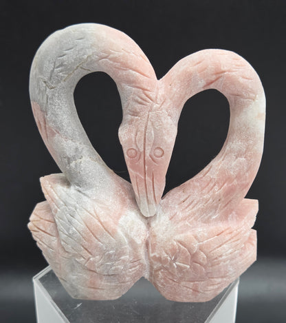A close-up of a sculpted decorative object featuring two intertwined looped forms resembling a stylized bird or abstract sculpture, mounted on a clear acrylic stand against a dark background.