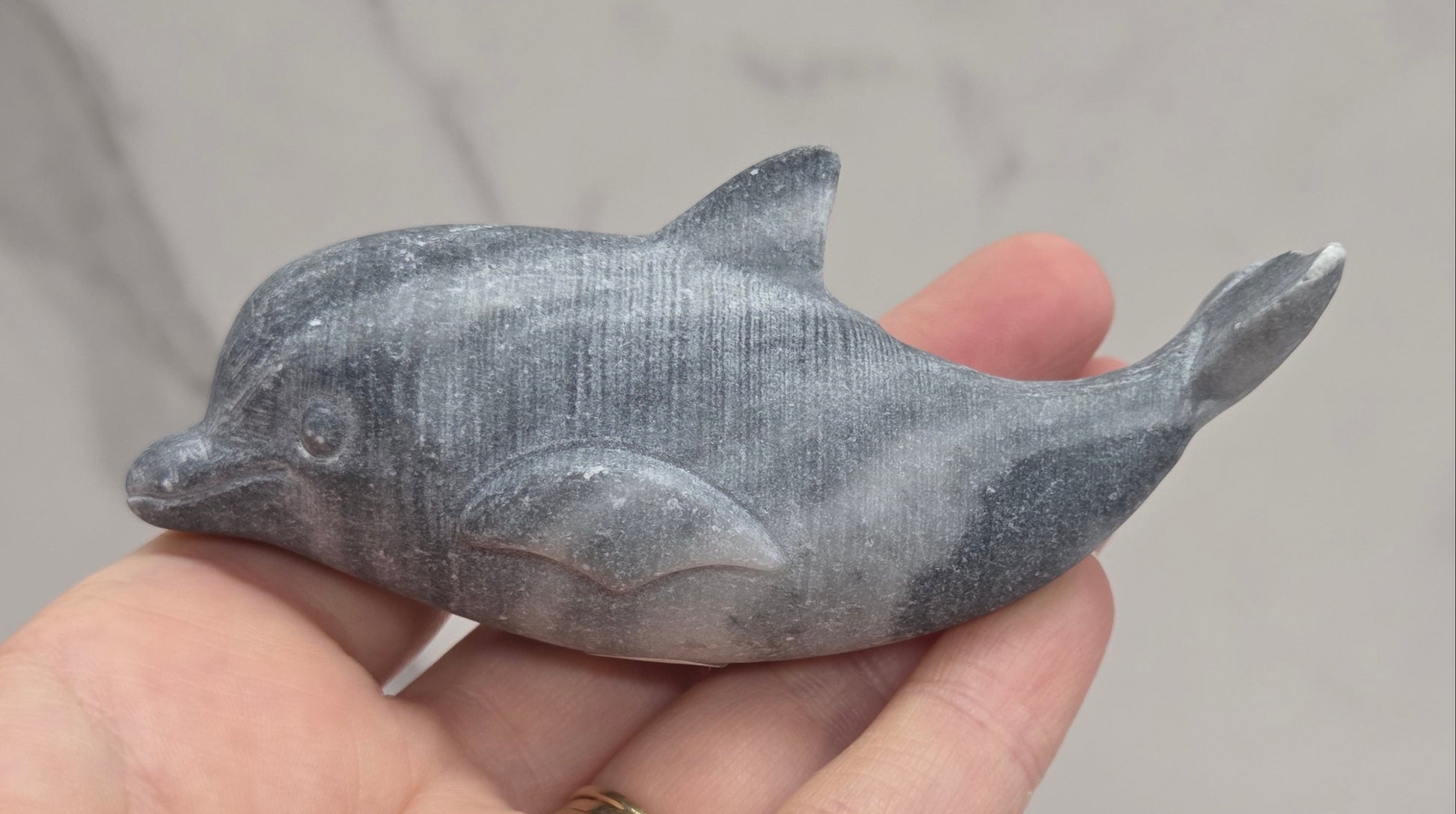 A small gray carved dolphin figurine held in a person's hand against a light gray marble-patterned background.
