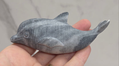 A small gray carved dolphin figurine held in a person's hand against a light gray marble-patterned background.