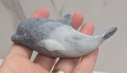 A hand holding a small gray stone dolphin figurine against a light gray marble-patterned background.