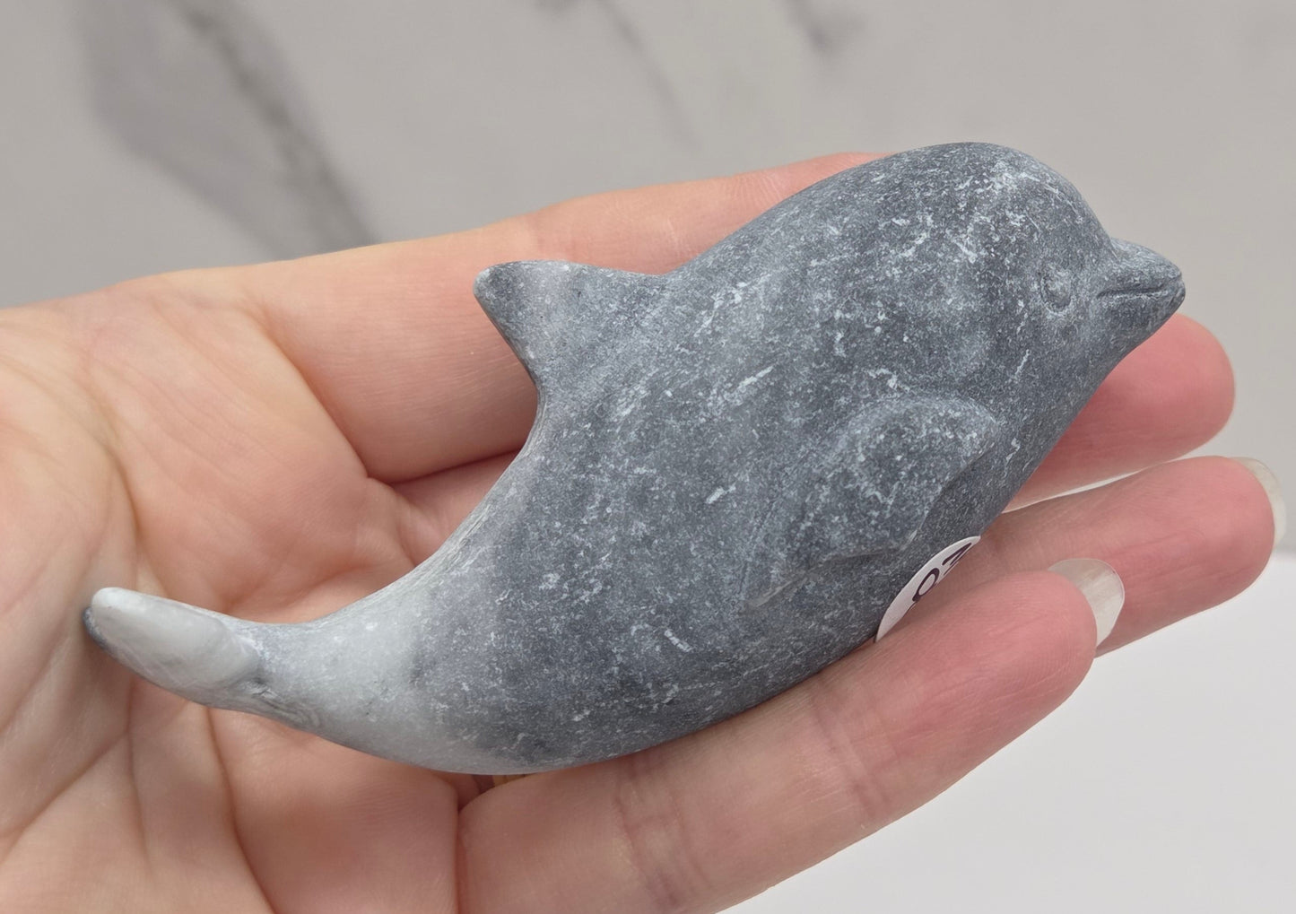 A small gray stone dolphin figurine held in a person's hand against a light marble-patterned background.