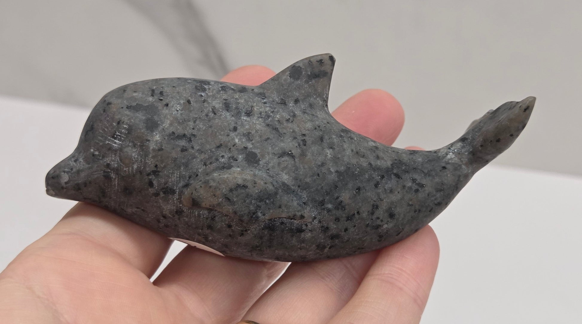 A hand holding a small carved dolphin figurine made of dark gray speckled stone, with a gold ring on the finger, against a light gray marble-patterned background.