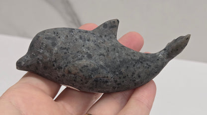 A hand holding a small carved dolphin figurine made of dark gray speckled stone, with a gold ring on the finger, against a light gray marble-patterned background.