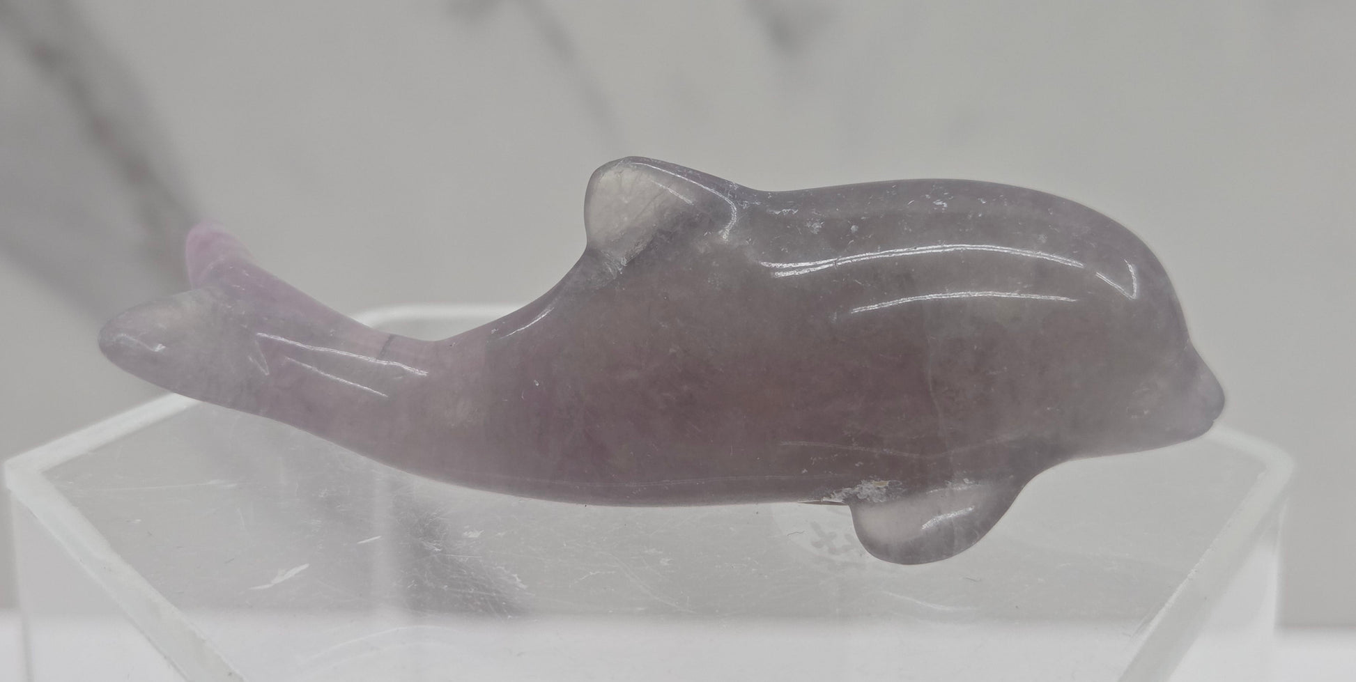 A polished carved dolphin figurine made of translucent lavender-purple stone, displayed on a clear acrylic stand against a light gray marble-patterned background.