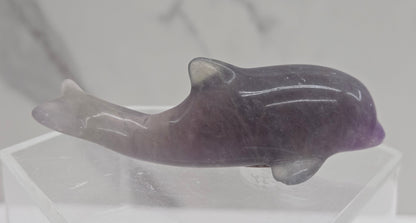 A polished carved dolphin figurine made of purple and gray stone, displayed on a clear acrylic stand against a light gray marble-patterned background.