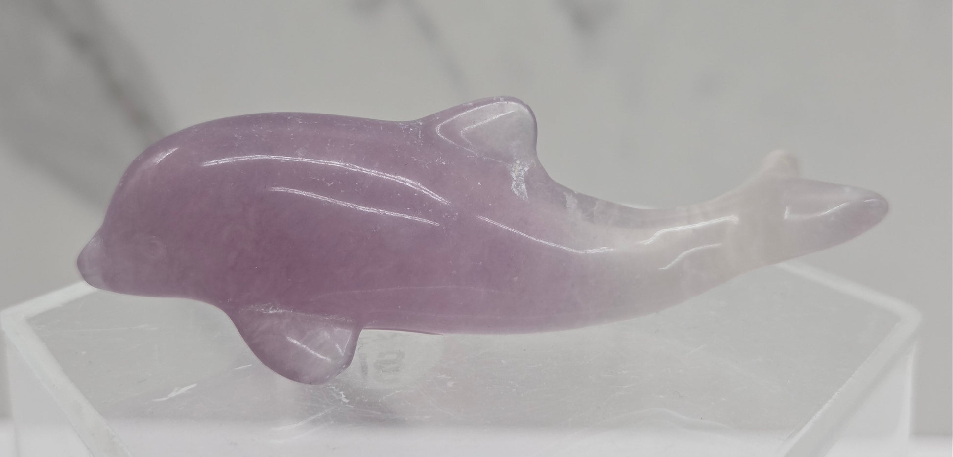 A polished pinkish-purple fluorite dolphin figurine displayed on a clear acrylic stand against a light gray marble-patterned background.