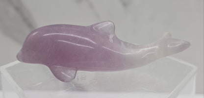 A polished pinkish-purple fluorite dolphin figurine displayed on a clear acrylic stand against a light gray marble-patterned background.