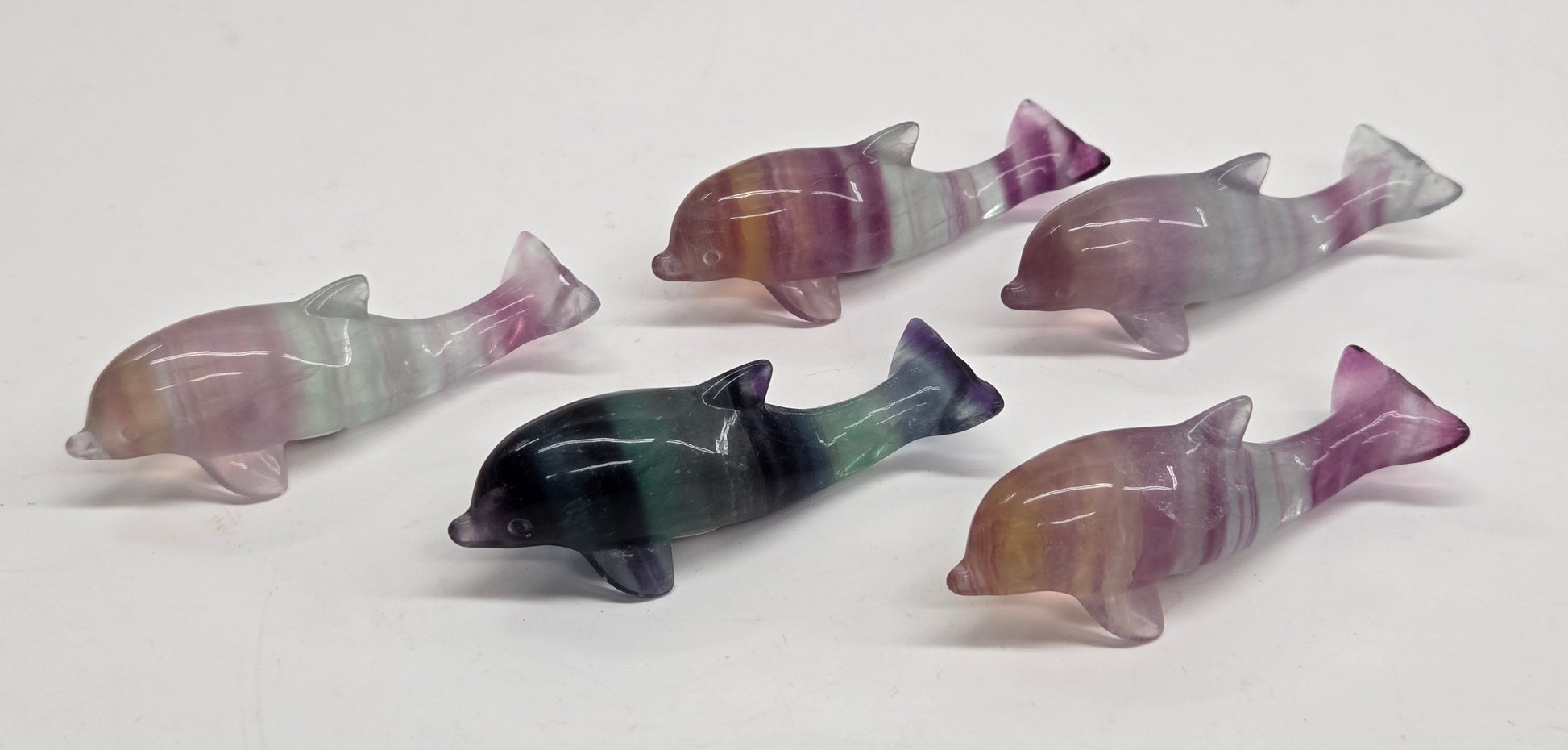 Five small polished dolphin figurines made from striped fluorite, displayed on a plain white surface. The dolphins feature natural banding in shades of purple, pink, green, and gray.