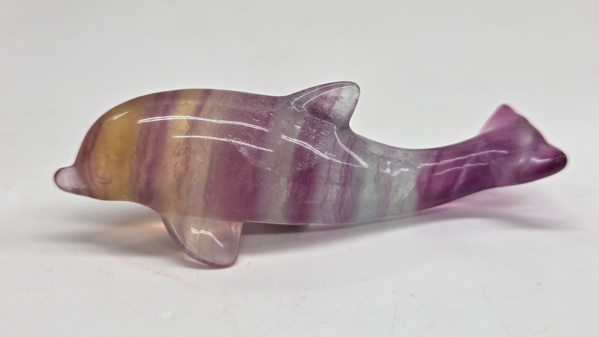 A polished natural fluorite dolphin figurine with purple, pink, and yellow banding, shown on a plain light gray background.