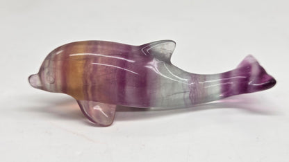 A polished dolphin figurine carved from purple and brown flúorite with white veining, displayed on a plain light gray background.