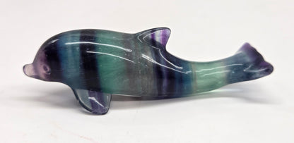 A polished dolphin figurine carved from fluorite, featuring a marbled pattern of green, blue, and purple, set against a plain white background.