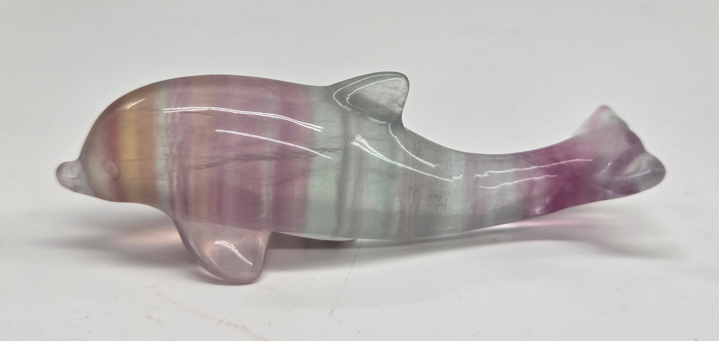 A polished stone dolphin figurine with vertical bands of purple, pink, and pale green, displayed on a plain light gray surface.