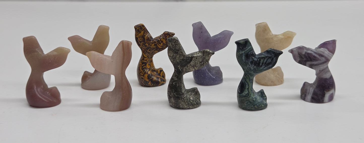 A set of eight small carved stone mermaid tail figurines in various earthy and purple tones, arranged in a row on a white surface with a light gray marble-patterned background.