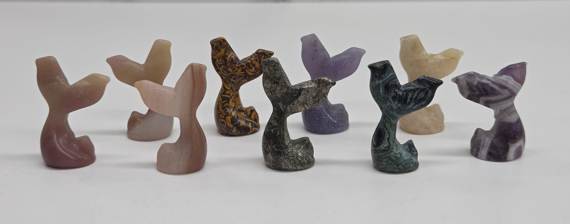 A set of eight small carved stone mermaid tail figurines in various earthy and purple tones, arranged in a row on a white surface with a light gray marble-patterned background.