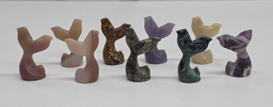 A set of eight small carved stone mermaid tail figurines in various earthy and purple tones, arranged in a row on a white surface with a light gray marble-patterned background.