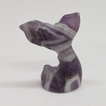 A small carved stone figurine shaped like a mermaid tail, made from dream amethyst, photographed against a plain light background.