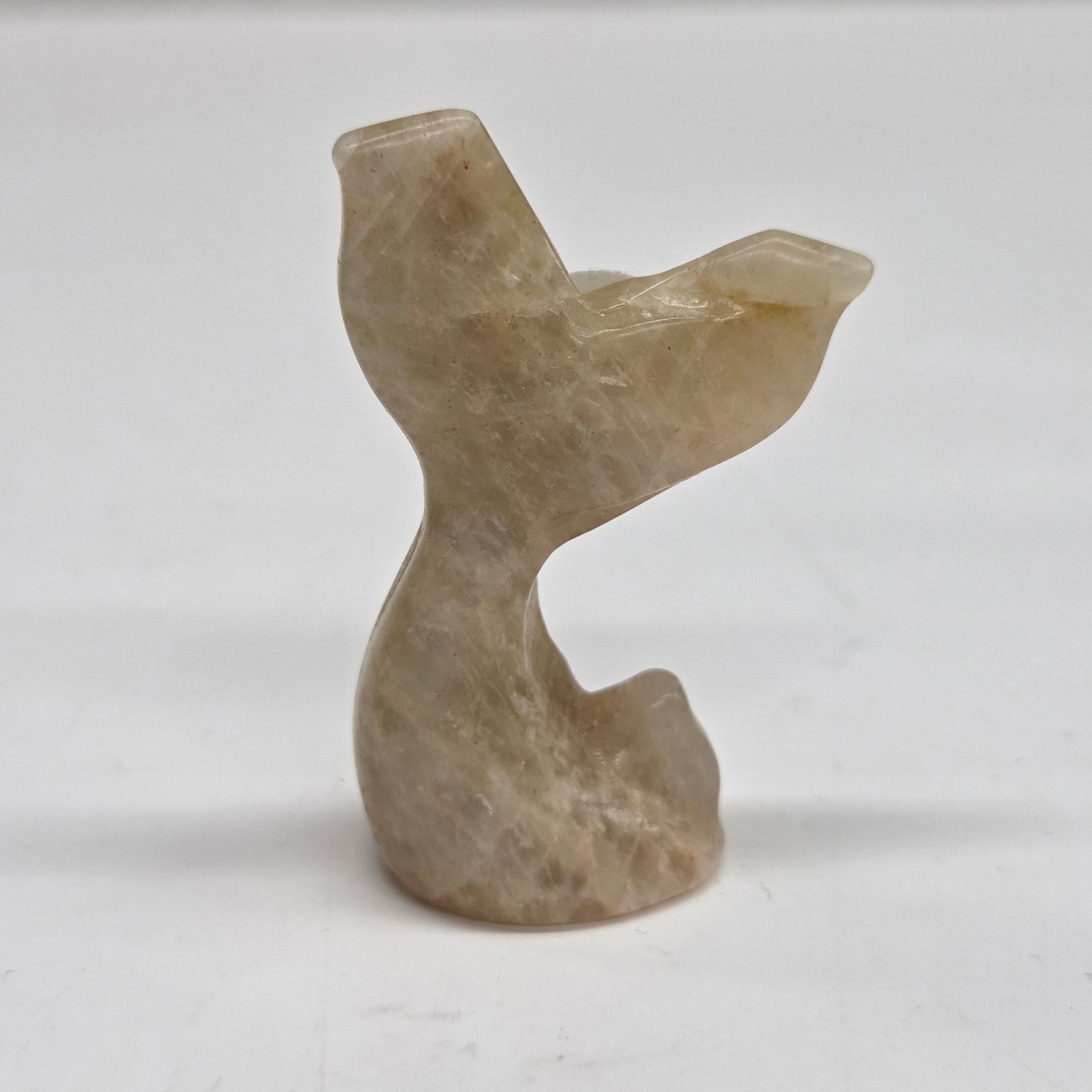 A small carved stone figurine shaped like a mermaid tail, made from moonstone, photographed against a plain light background.