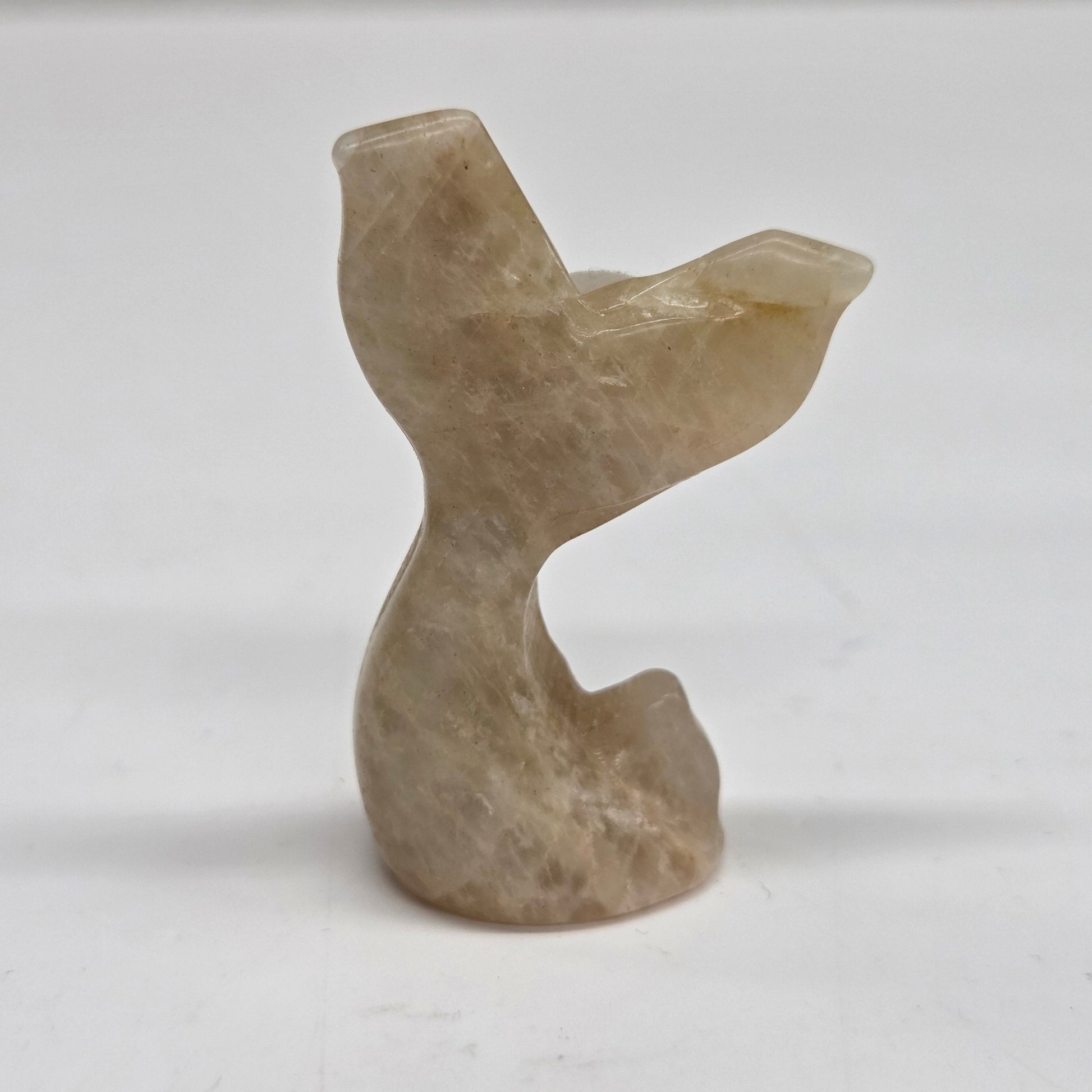 A small carved stone figurine shaped like a mermaid tail, made from moonstone, photographed against a plain light background.
