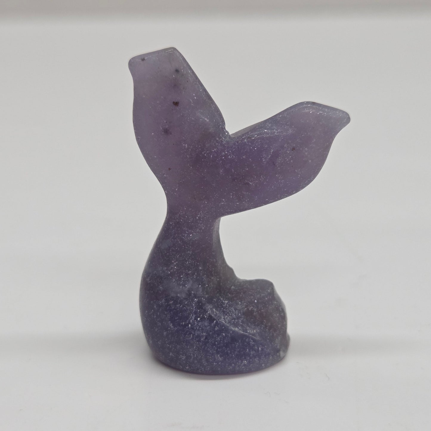 A small carved stone figurine shaped like a mermaid tail, made from lepidolite, photographed against a plain light background.
