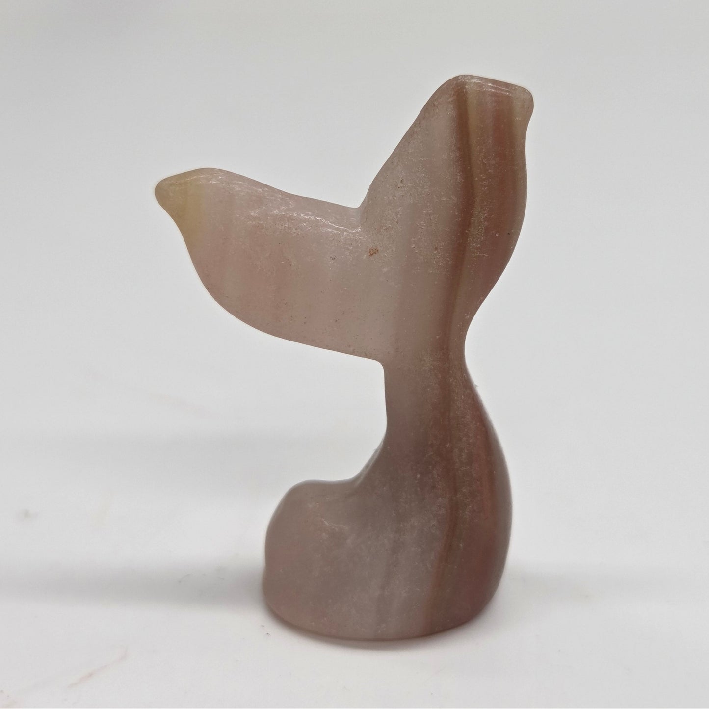 A small carved stone figurine shaped like a mermaid tail, made from banded pink calcite photographed against a plain light background.