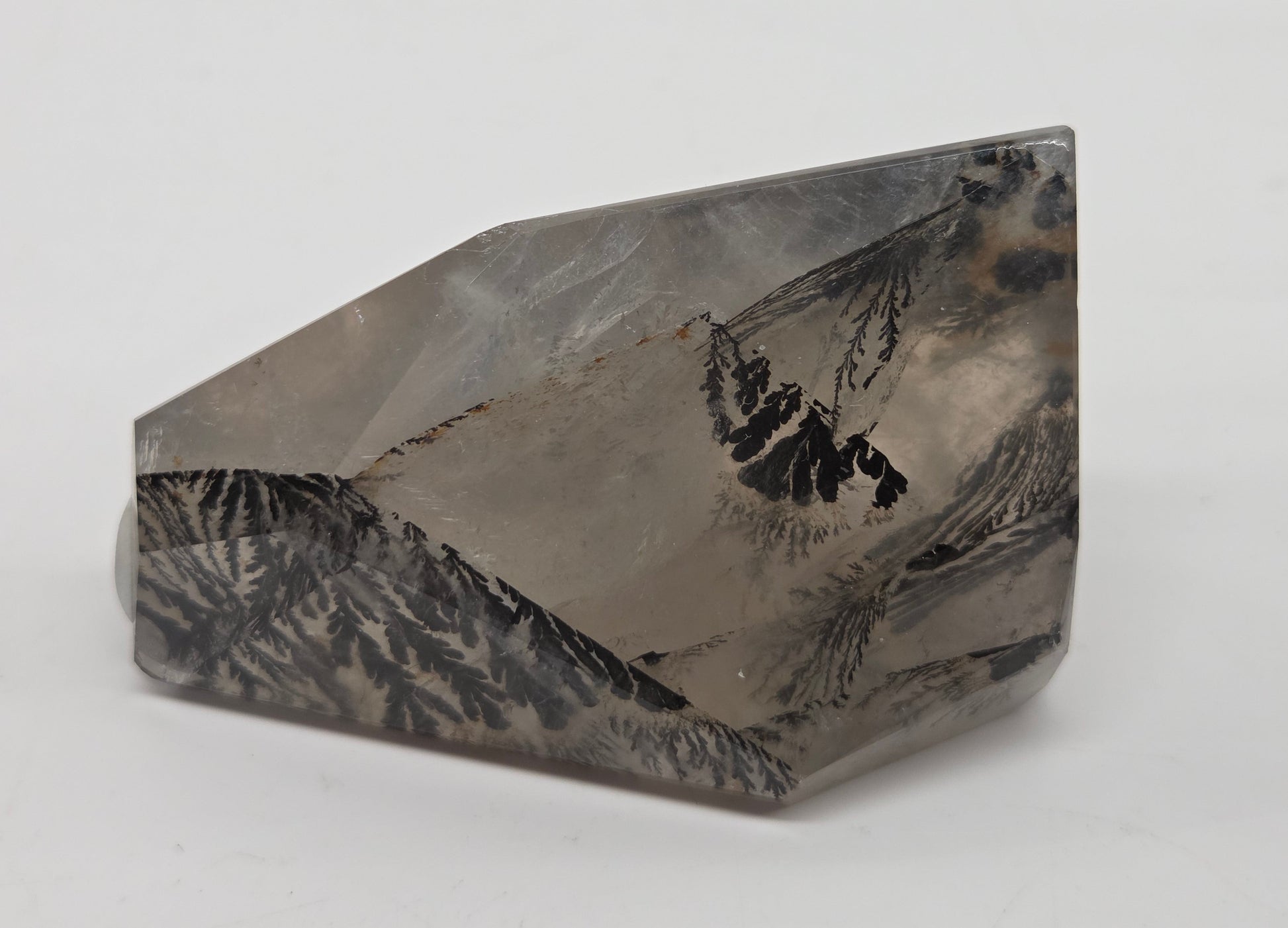 A faceted crystal mineral specimen with a mountain landscape pattern, featuring gray, black, and white tones, set against a plain light gray background.