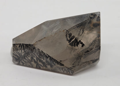 A faceted crystal mineral specimen with a mountain landscape pattern, featuring gray, black, and white tones, set against a plain light gray background.