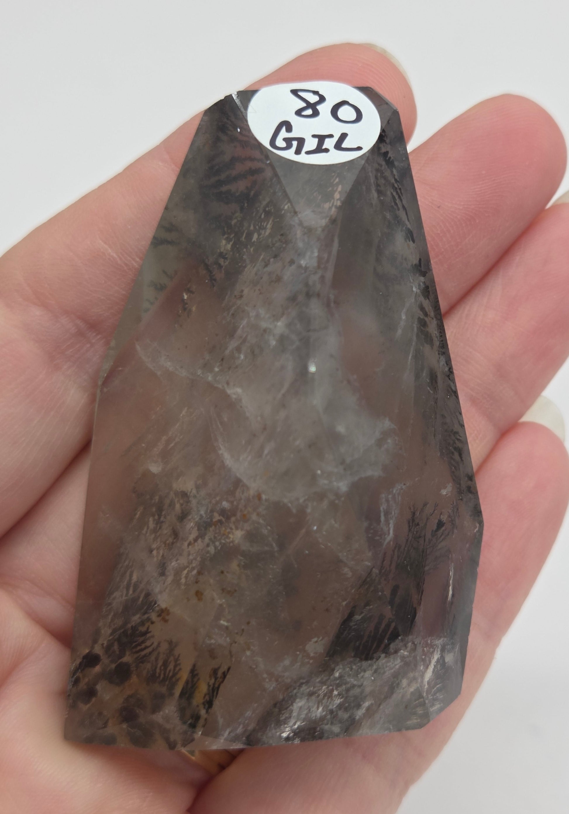 A hand holding a raw smoky quartz crystal point against a plain white background, with a small white label reading '80 GIL' attached near the tip.