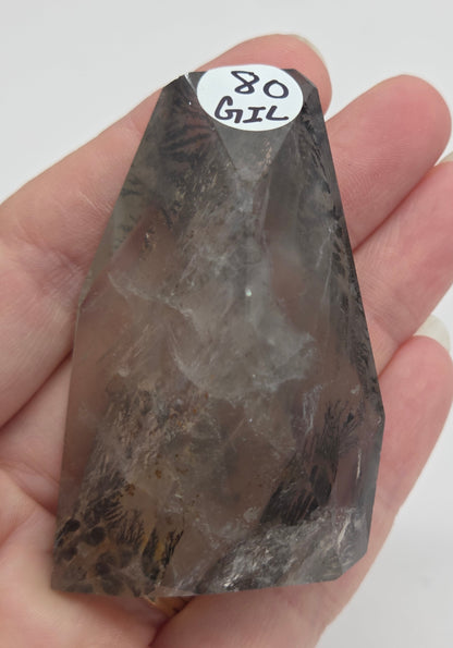 A hand holding a raw smoky quartz crystal point against a plain white background, with a small white label reading '80 GIL' attached near the tip.