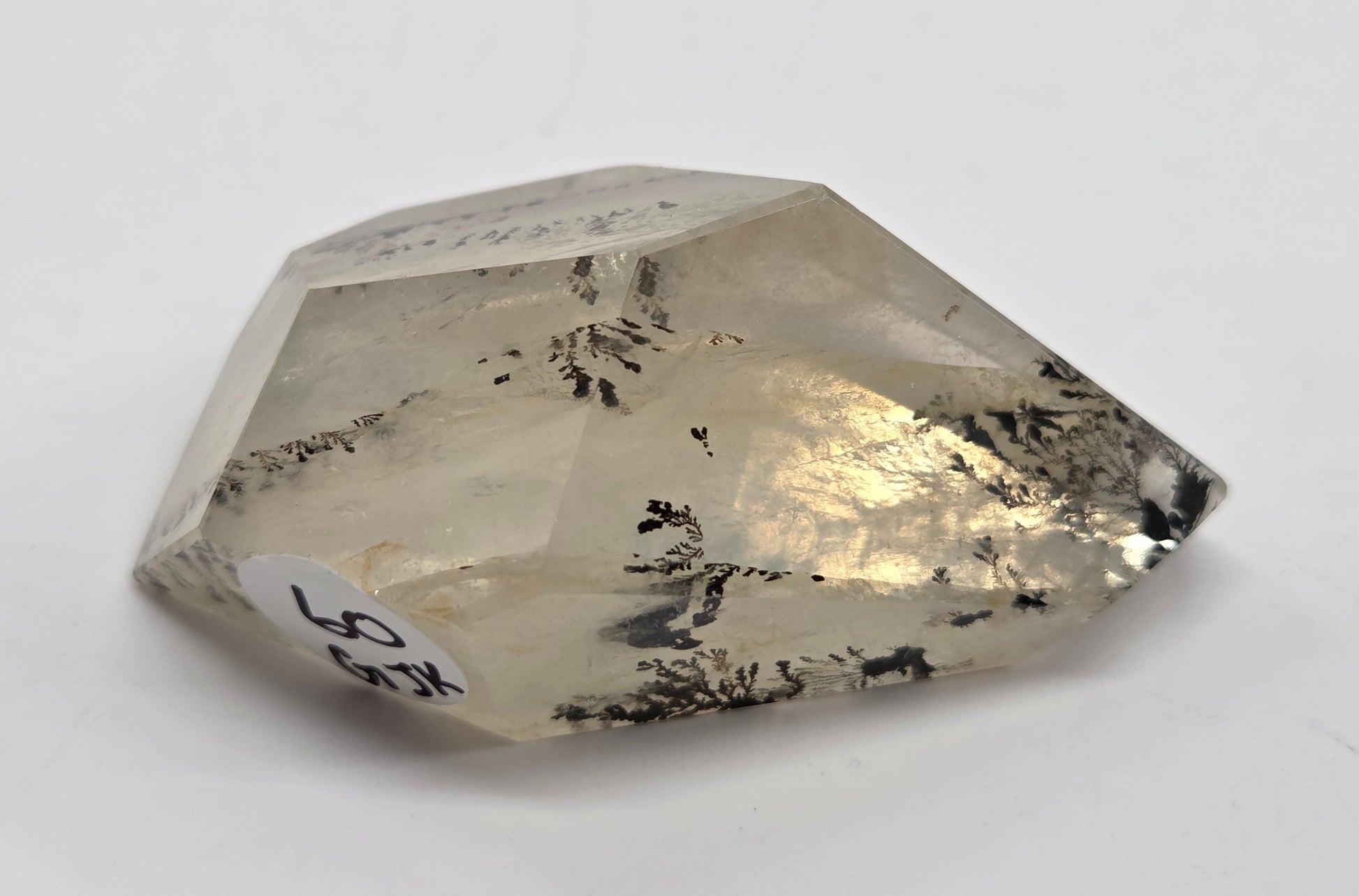 A faceted clear crystal gemstone with black inclusions, photographed on a plain light background. A small white label with handwritten text is attached near one corner.