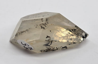 A faceted clear crystal gemstone with black inclusions, photographed on a plain light background. A small white label with handwritten text is attached near one corner.