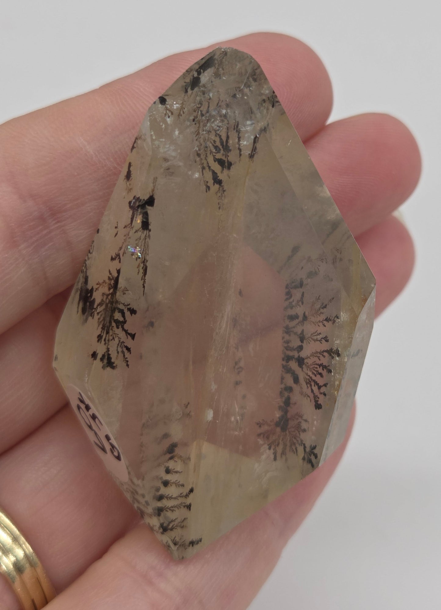 A hand holding a raw, translucent pale yellow crystal with dark inclusions, against a plain light gray background.
