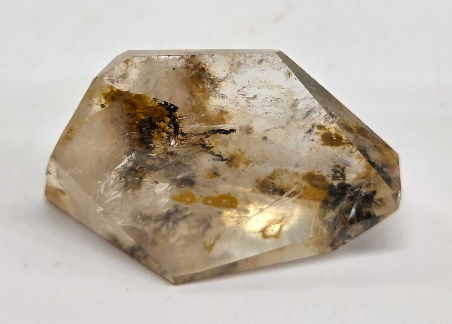 A faceted raw crystal stone with a translucent off-white body, containing natural inclusions and dark brown to black inclusions, photographed on a plain light background.