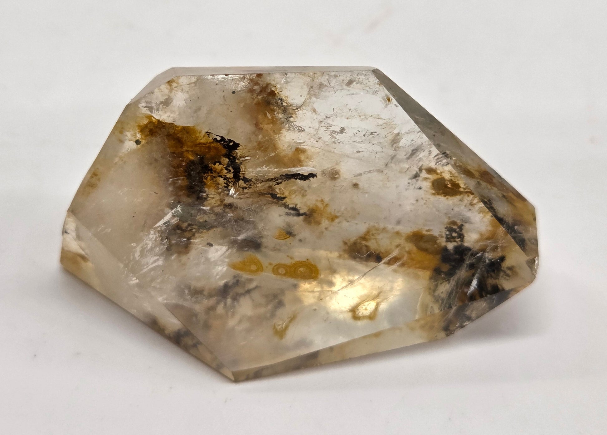 A faceted raw crystal stone with a translucent off-white body, containing natural inclusions and dark brown to black inclusions, photographed on a plain light background.
