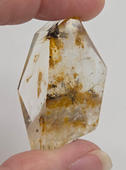 A person's hands holding a clear faceted crystal point with natural golden-brown and dark inclusions, shown against a plain light background.
