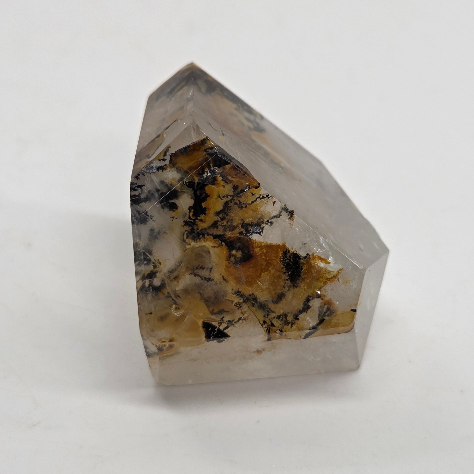 A faceted clear crystal point with natural brown, black, and white inclusions, photographed on a plain light gray background.