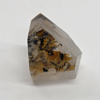 A faceted clear crystal point with natural brown, black, and white inclusions, photographed on a plain light gray background.