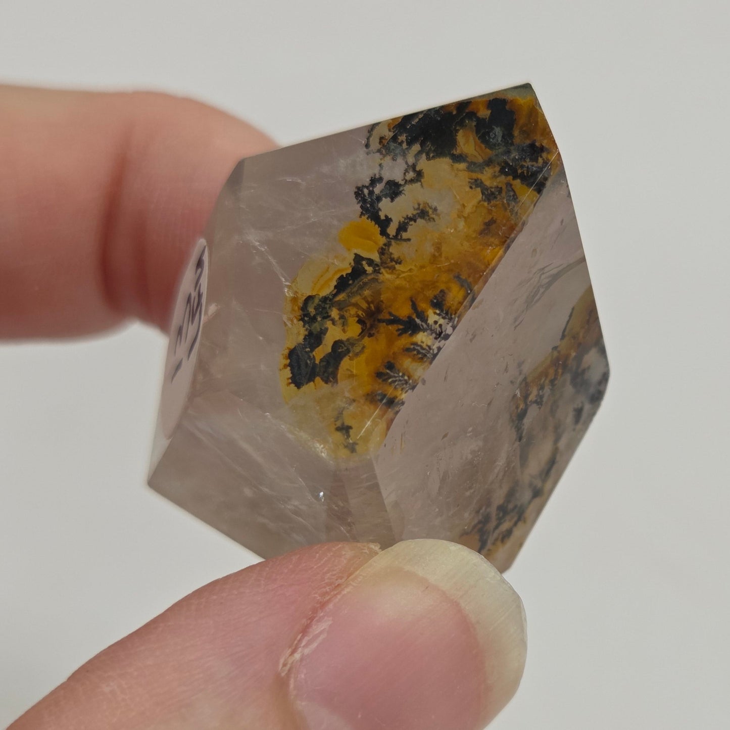 A hand holding a small faceted clear crystal point with yellow and dark brown inclusions, shown against a plain light background.