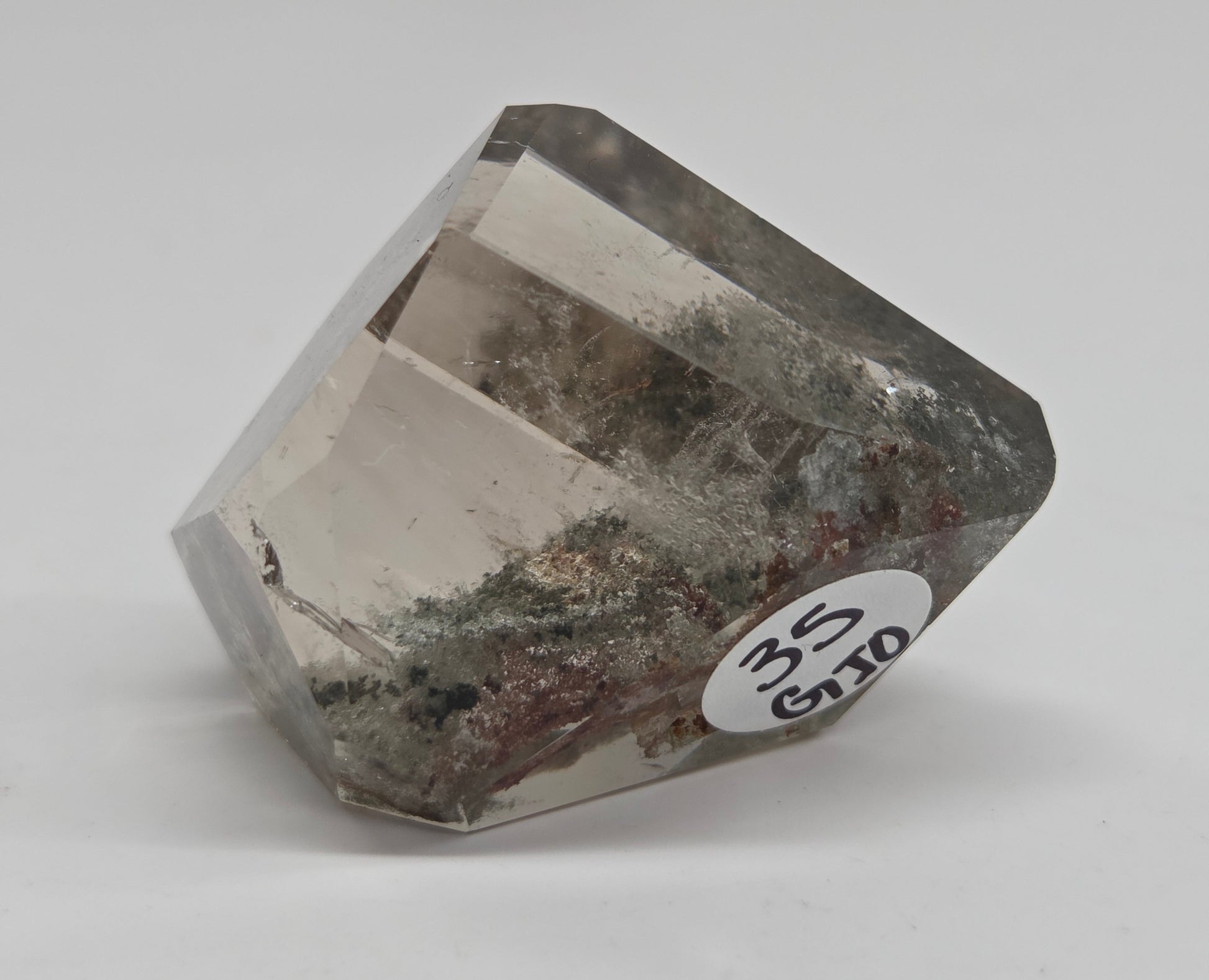 A faceted clear gemstone crystal with natural inclusions and a white circular label reading '3.5 CT' on a light gray background.