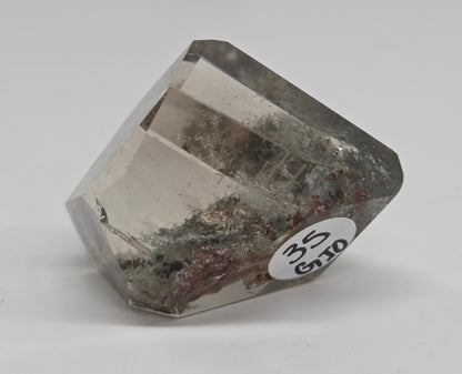 A faceted clear gemstone crystal with natural inclusions and a white circular label reading '3.5 CT' on a light gray background.