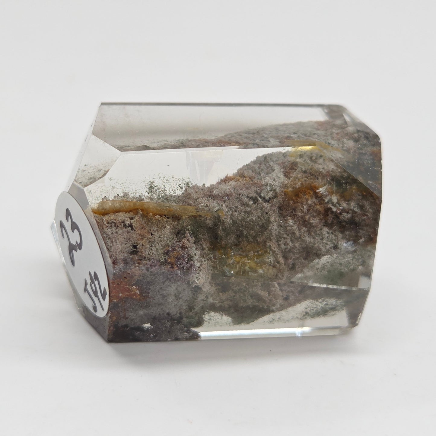 A faceted clear crystal or gemstone with inclusions and a mottled brown, gray, and gold interior, photographed on a plain light background. A small white label with black numbers is attached to one side.