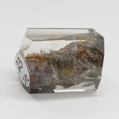 A faceted clear crystal or gemstone with inclusions and a mottled brown, gray, and gold interior, photographed on a plain light background. A small white label with black numbers is attached to one side.