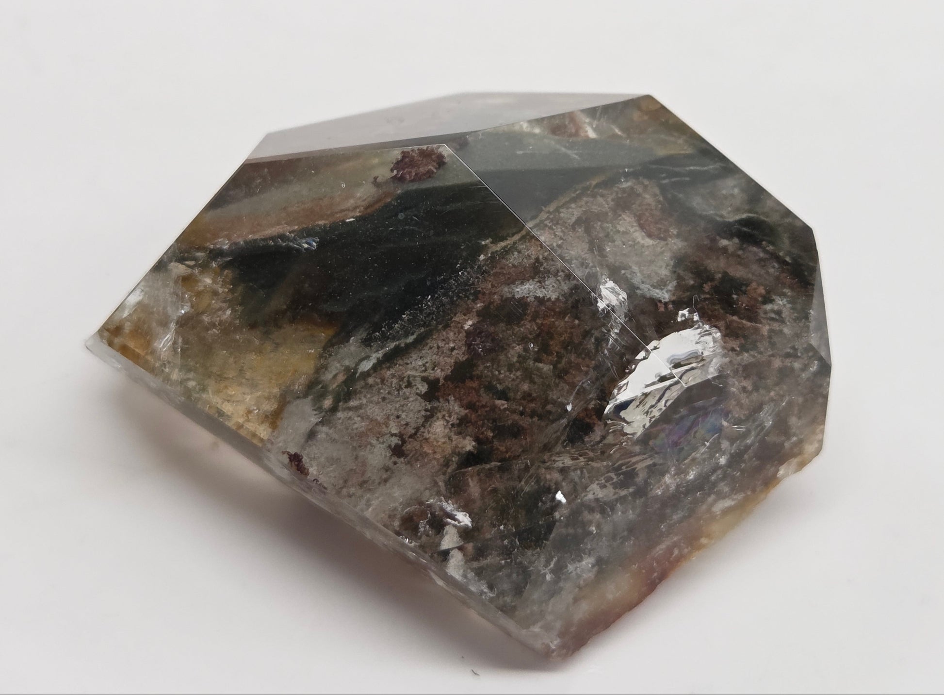A faceted crystal gemstone with a natural earthy pattern, featuring shades of dark brown, black, gray, and subtle beige highlights, set against a plain white background.