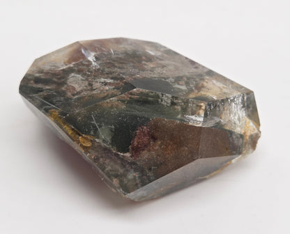 A faceted raw gemstone with a dark gray and black base, showing natural inclusions and internal brown and reddish-brown pockets, photographed on a plain white background.