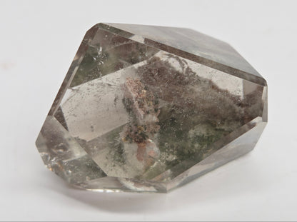 A faceted clear gemstone with natural inclusions and smoky gray internal areas, photographed on a plain light background.