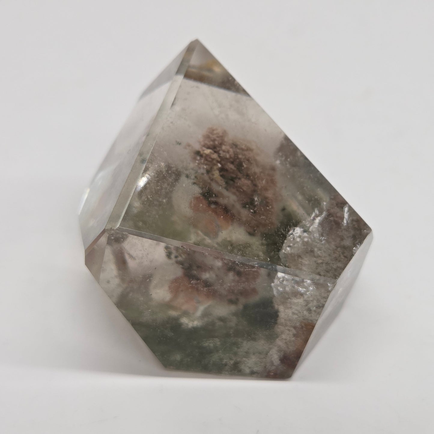 A faceted clear crystal or glass-like geometric ornament with internal cloudy inclusions and a small brownish textured area near the center, photographed on a plain light background.