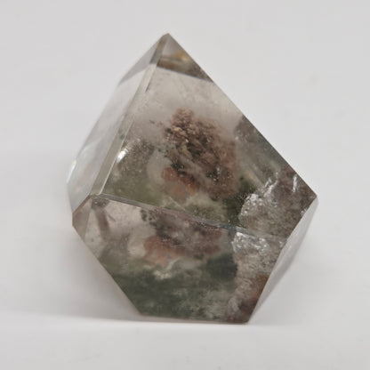 A faceted clear crystal or glass-like geometric ornament with internal cloudy inclusions and a small brownish textured area near the center, photographed on a plain light background.