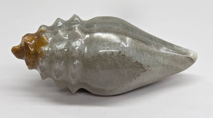 A polished gray stone shell sculpture with a textured spiral form and a warm brown accent at the pointed spiral end, displayed on a plain white surface.