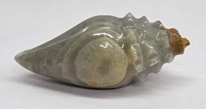 A polished stone seashell sculpture with a smooth, glossy finish, featuring a muted gray-green body with warm beige and brown highlights, set against a plain white background.