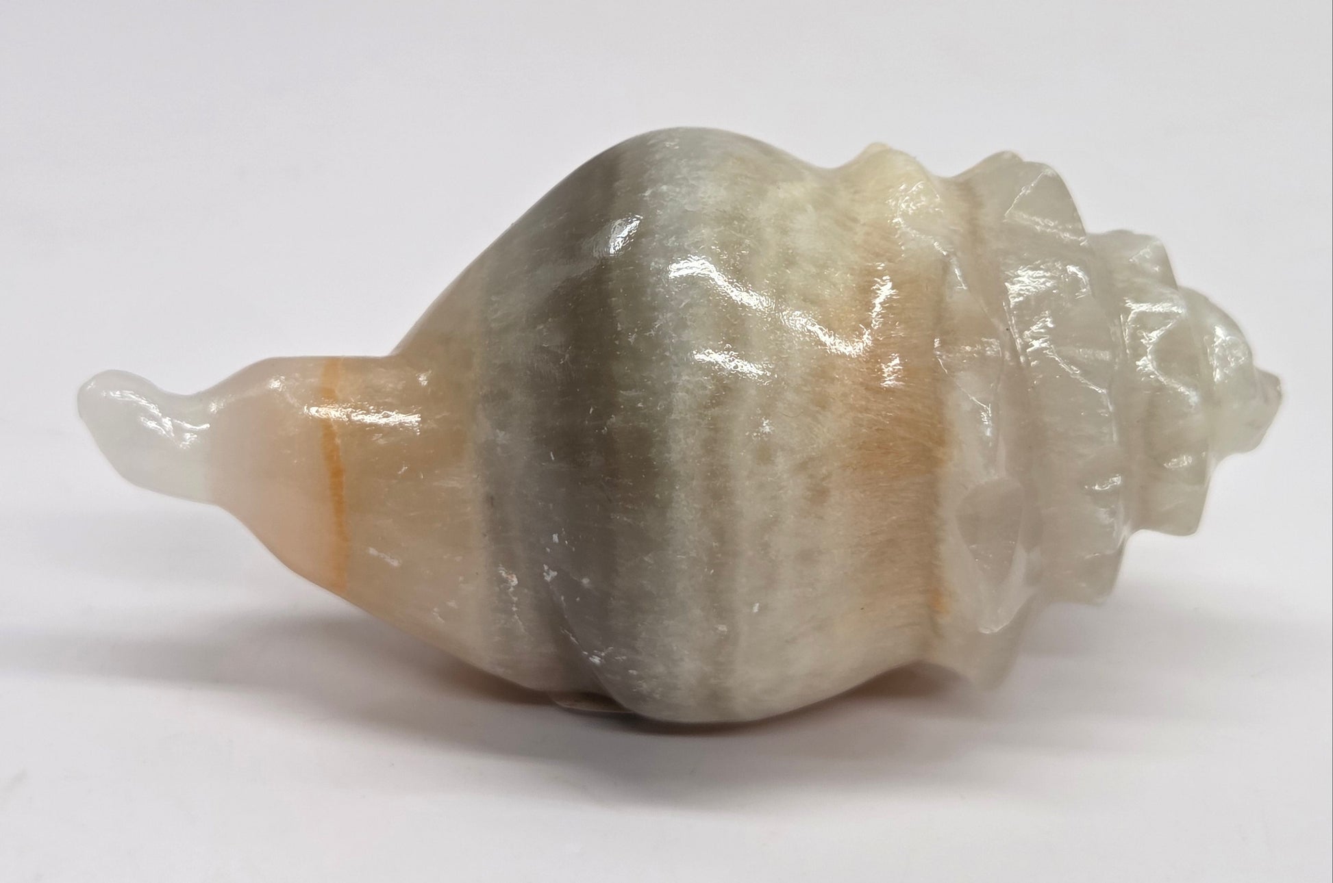 A carved stone shell sculpture with a spiral seashell shape, featuring a smooth, polished finish in shades of gray, beige, and white, set against a plain light gray background.